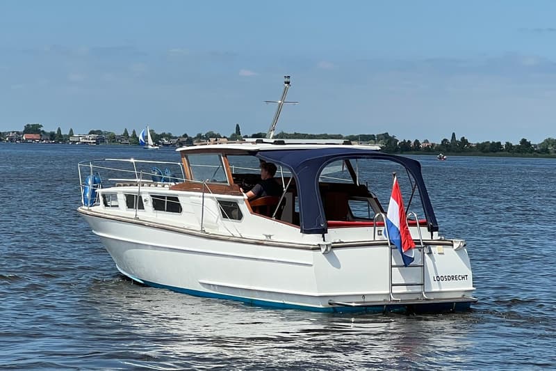 Een witte motorboot met blauwe kap en Nederlandse vlag vaart op het water, met "LOOSDRECHT" op de spiegel.
