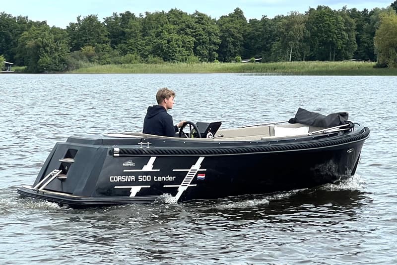 Een zwarte Corsiva 500 tender vaart op het water met een persoon aan het stuur, tegen een achtergrond van bomen.