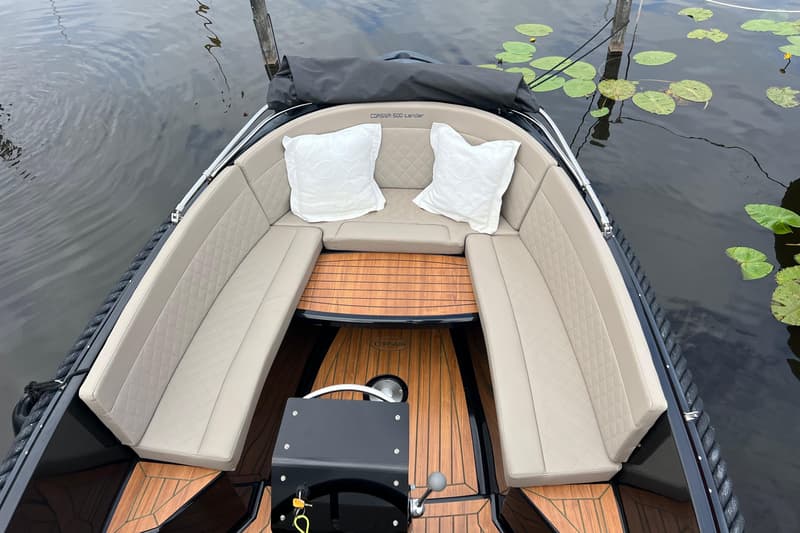 Een bovenaanzicht van een boot met beige kussens, een houten vloer en een zwart stuur, liggend in het water.