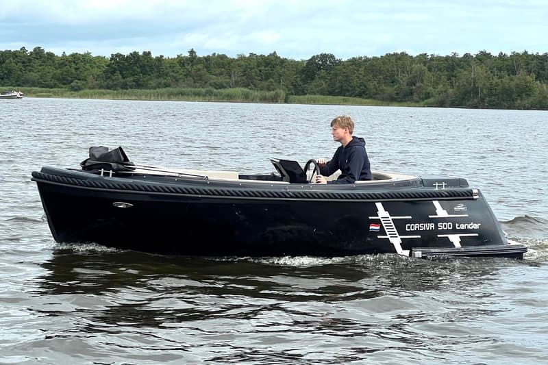 Een jongeman bestuurt een zwarte Corsiva 500 tender op het water, met een bosrijke oever op de achtergrond.