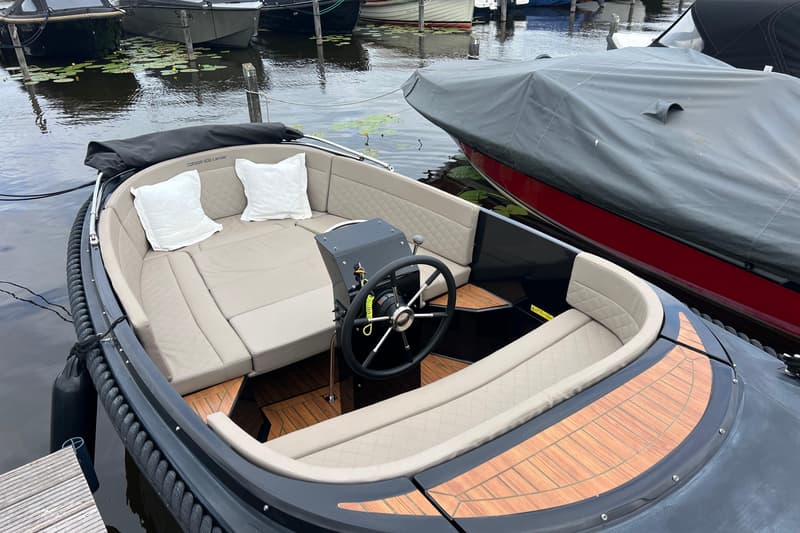 Een donkergrijze sloep met beige kussens en een houten vloer ligt aangemeerd naast een rode boot met een grijs afdekzeil.