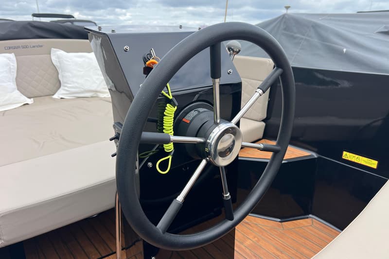 Een zwart stuurwiel met chromen spaken en een gele veiligheidslijn op een boot met beige kussens en een houten vloer.