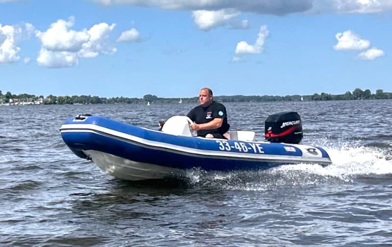 Een man vaart in een blauw-witte rubberboot met een zwarte Mercury buitenboordmotor over water onder een bewolkte hemel.