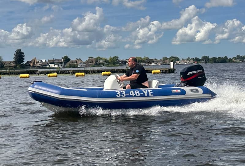Een man vaart in een blauwe rubberboot met een Mercury buitenboordmotor over water, met huizen op de achtergrond.