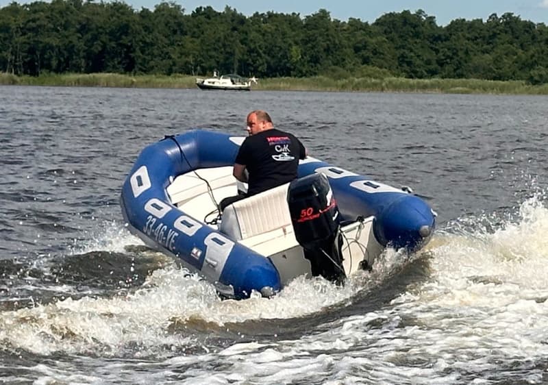 Een man in een blauw-witte rubberboot met een zwarte buitenboordmotor vaart over water, met een bosrijke oever op de…