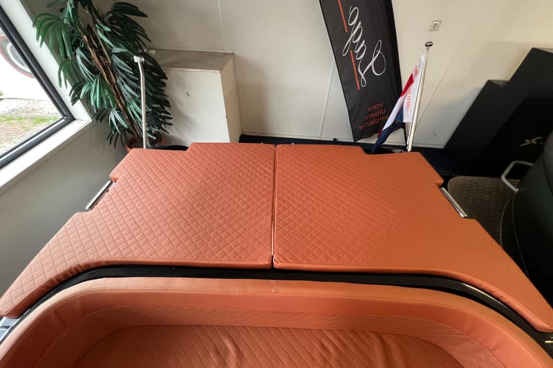 De foto toont een interieur met oranje, gestoffeerde zitplaatsen en een liggedeelte, een raam, een plant en een vlag.