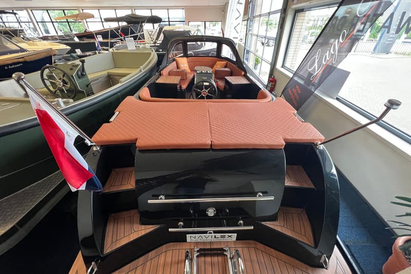 Een zwarte boot met bruine kussens en een Nederlandse vlag, staand in een showroom met andere boten. — Lago Amore 606 Tender