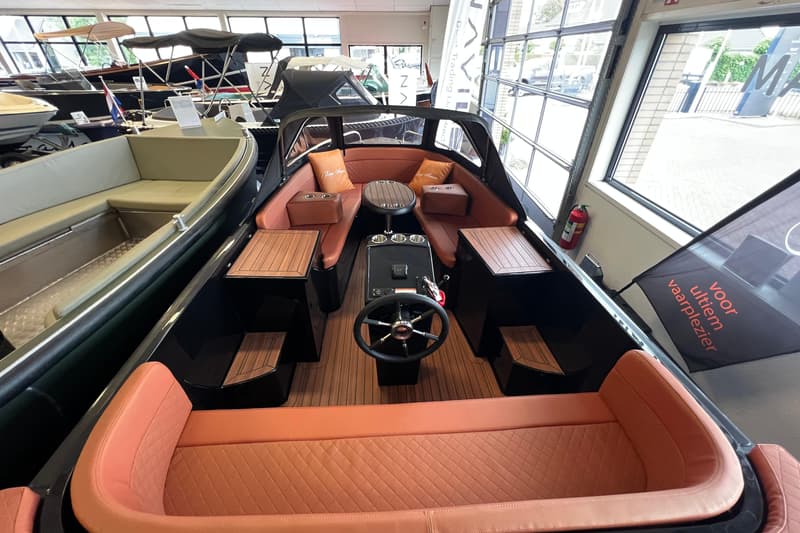 Een zwarte boot met oranje kussens en een houten vloer staat in een showroom met andere boten. — Lago Amore 606 Tender