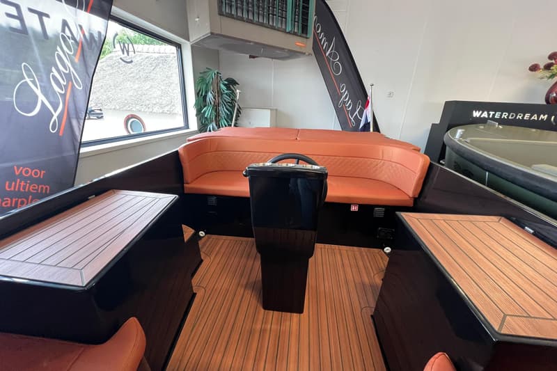 De foto toont het interieur van een boot met oranje gestoffeerde banken, een zwart stuurconsole en een houten vloer.