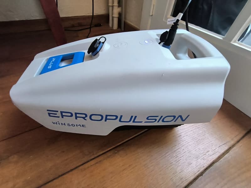 Een witte EPROPULSION WIN SOME accu met blauwe accenten en een blauw "PLUS" handvat, staand op een houten vloer.