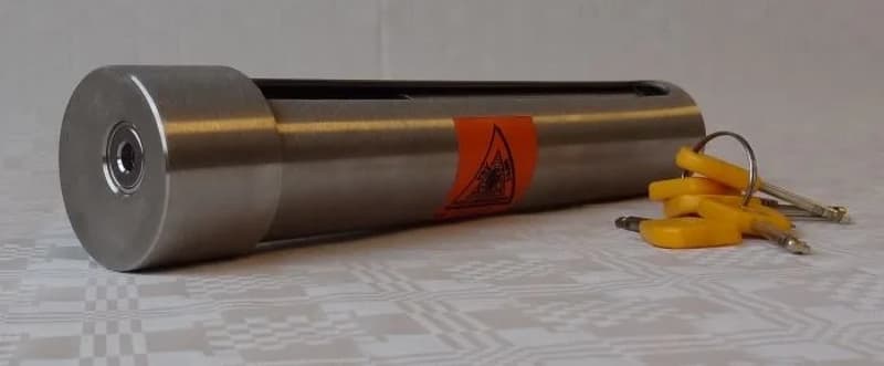 Een zilverkleurig, cilindervormig voorwerp met een oranje sticker en een sleutelgat aan één uiteinde, met een bosje…