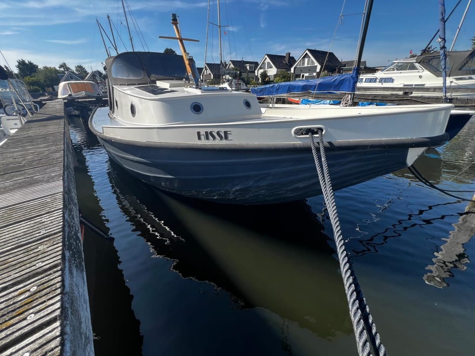 Een wit-grijze boot genaamd "HISSE" ligt afgemeerd aan een houten steiger, met een zwarte buiskap en ronde patrijspoorten.